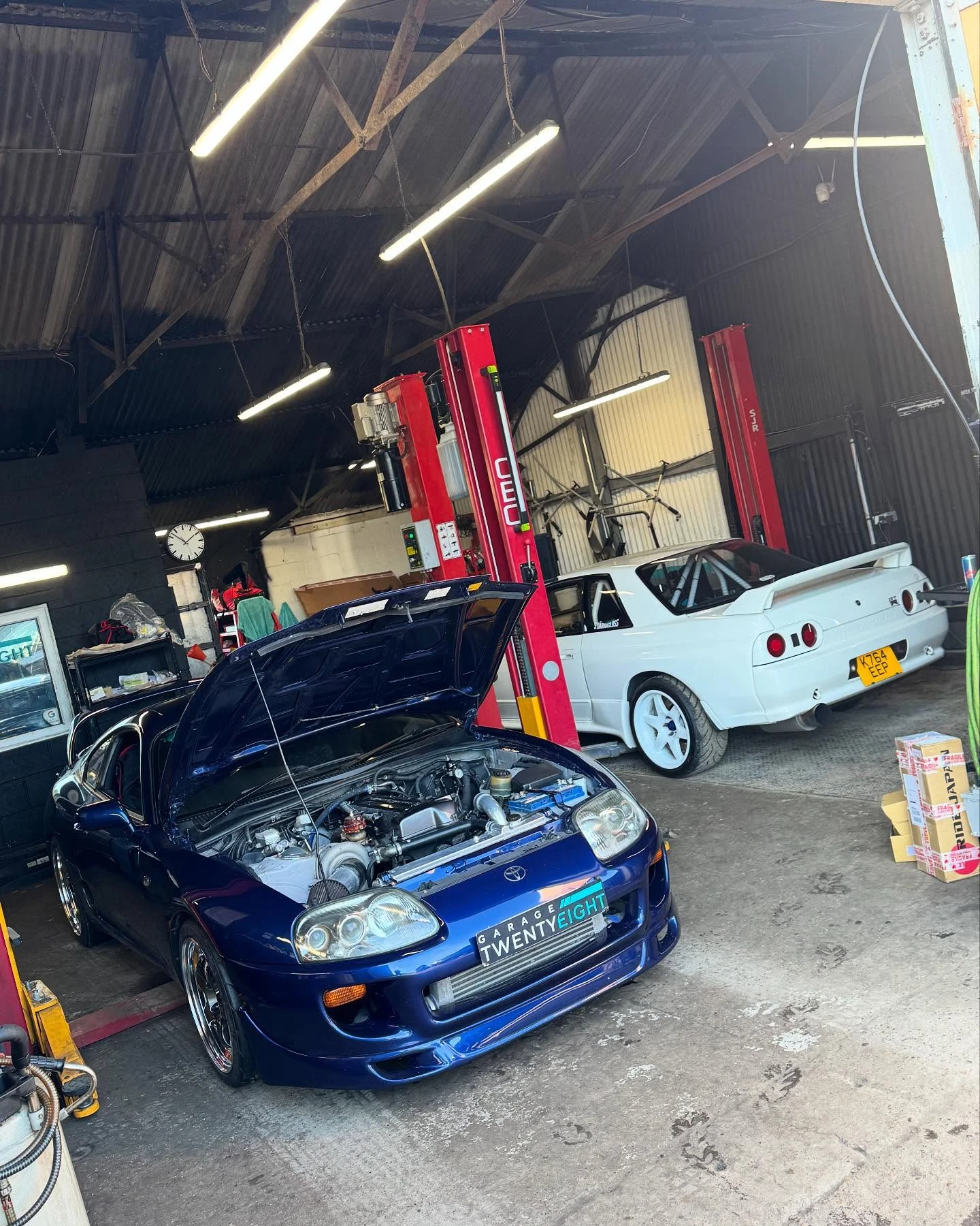 Blue Toyota Supra with open hood and white Nissan Skyline in a garage with red car lifts.