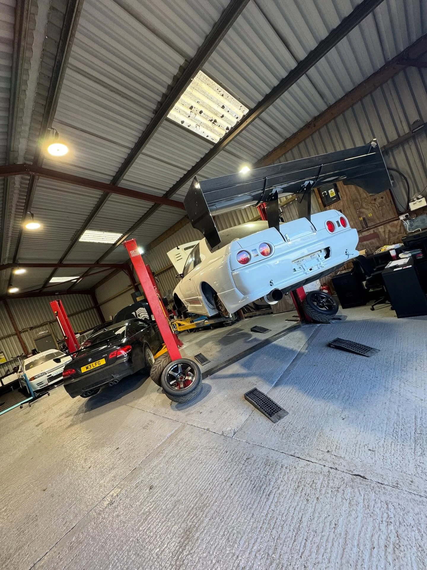 White sports car with large rear wing elevated on a lift inside a garage with other cars.