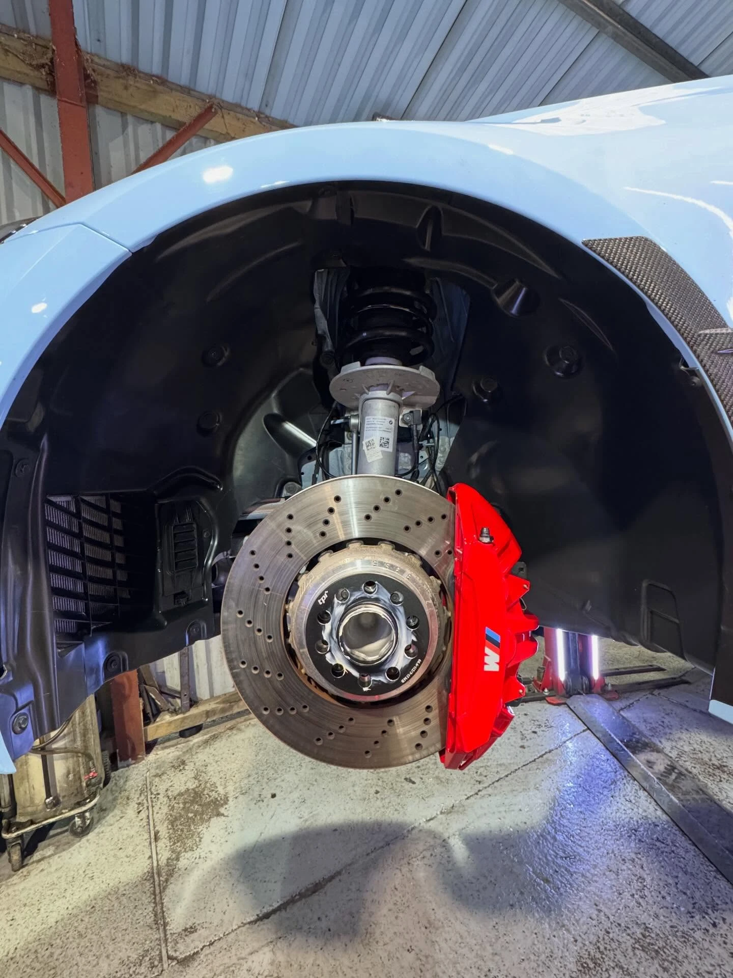 Close-up of a car's wheel hub with a drilled brake disc and red BMW M brake caliper in a garage.