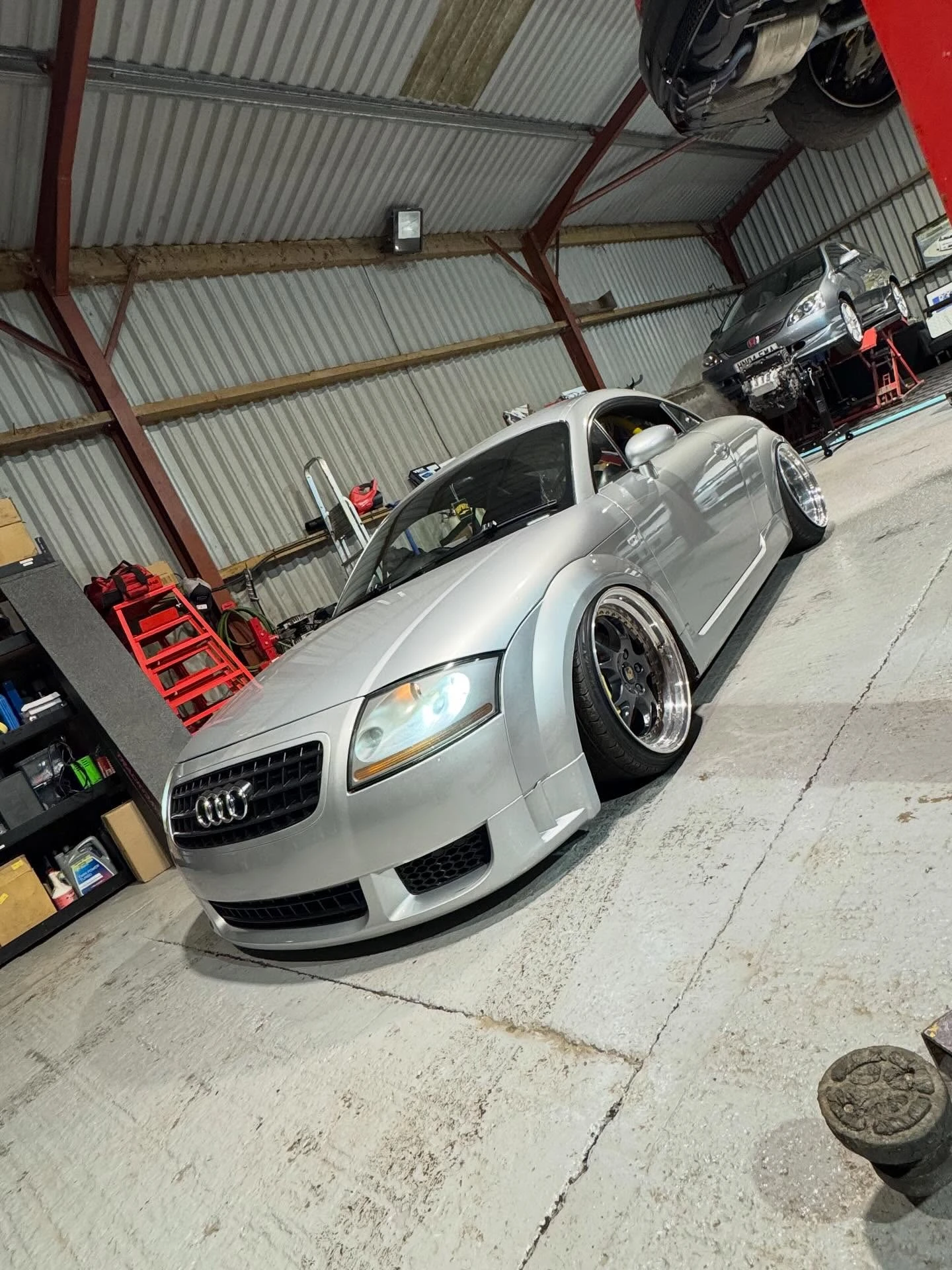 Silver Audi TT with lowered suspension and aftermarket wheels in a garage with tools and another car.