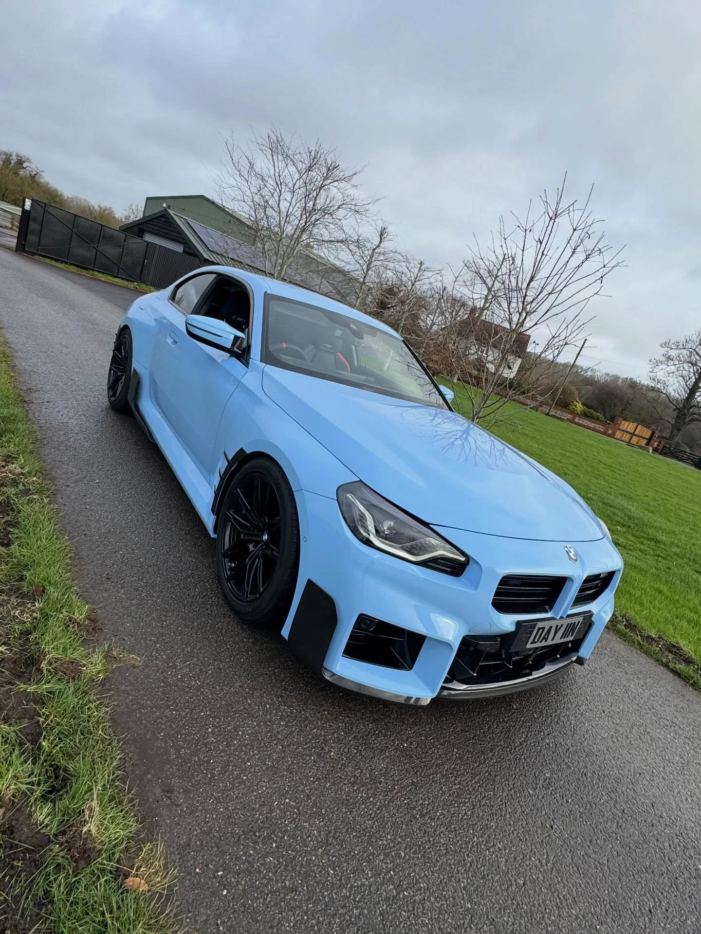 Light blue sporty BMW car parked on wet road with green grass and trees in background.
