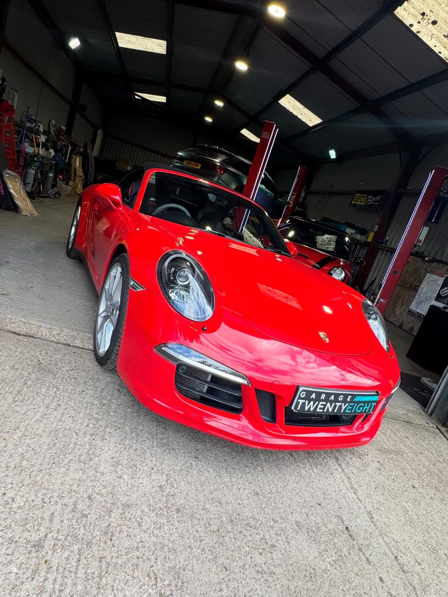 Red Porsche sports car parked inside a garage with tools and other cars in the background.