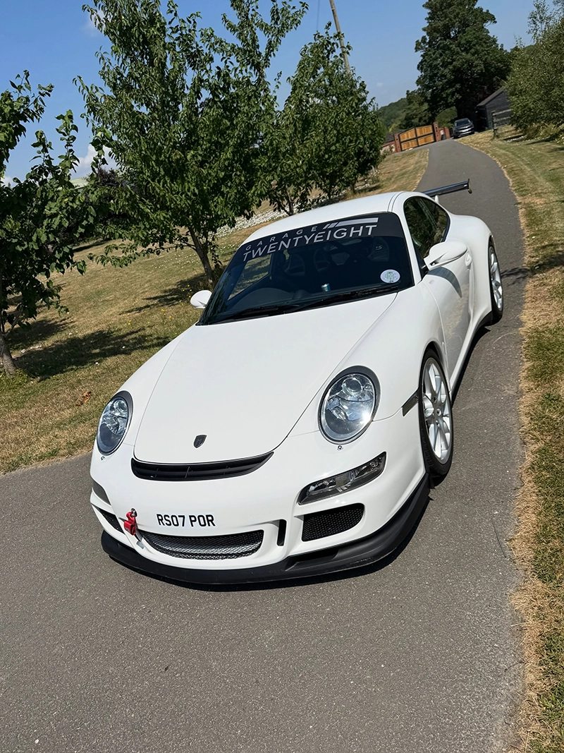 White sports car with spoiler parked on a narrow road lined with trees and grass.
