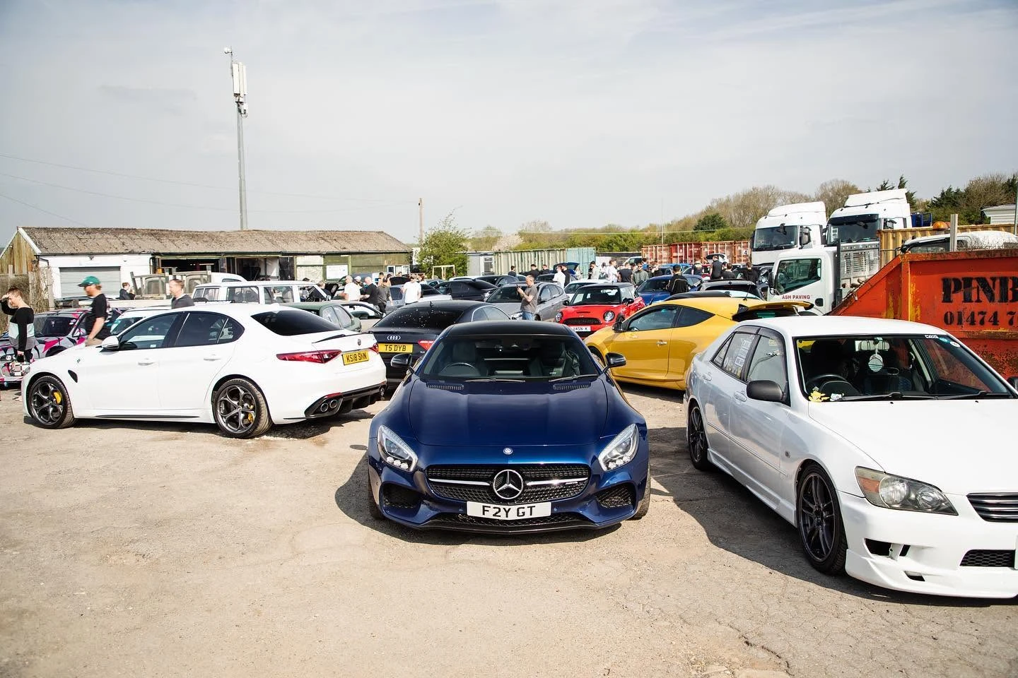 Blue Mercedes sports car parked among various cars and people at an outdoor car meet.