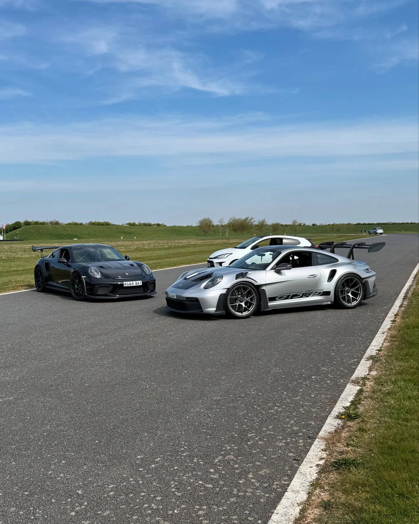 Two Porsche sports cars, black and silver GT3 RS, parked on an empty racetrack under a blue sky.