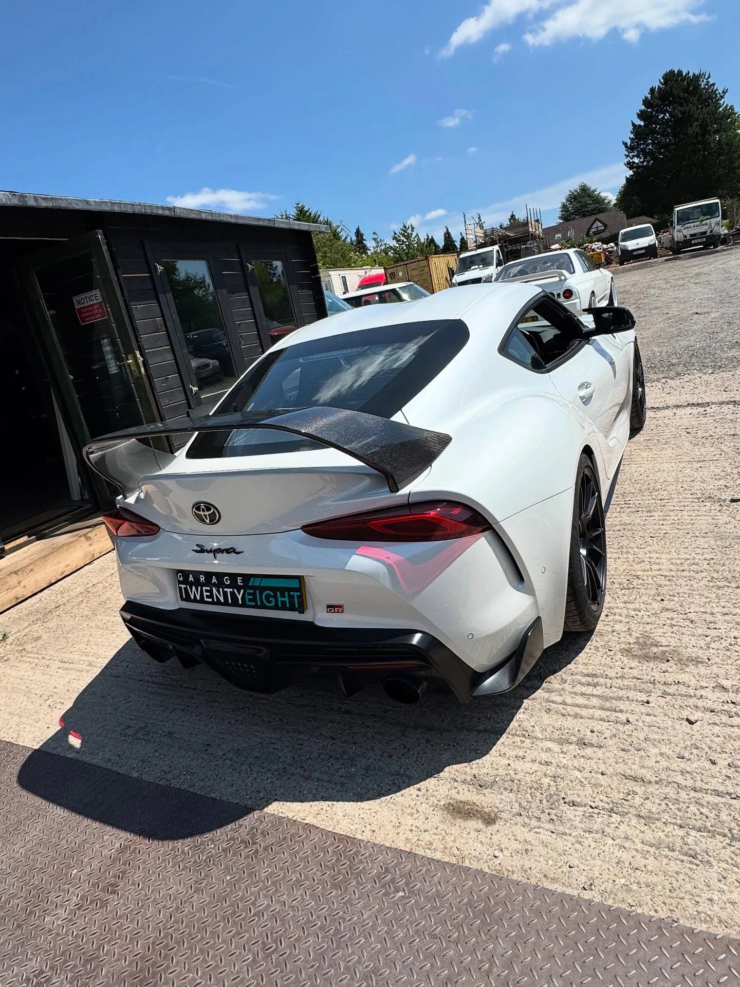 White Toyota Supra sports car with a large rear spoiler parked near a black building on a sunny day.
