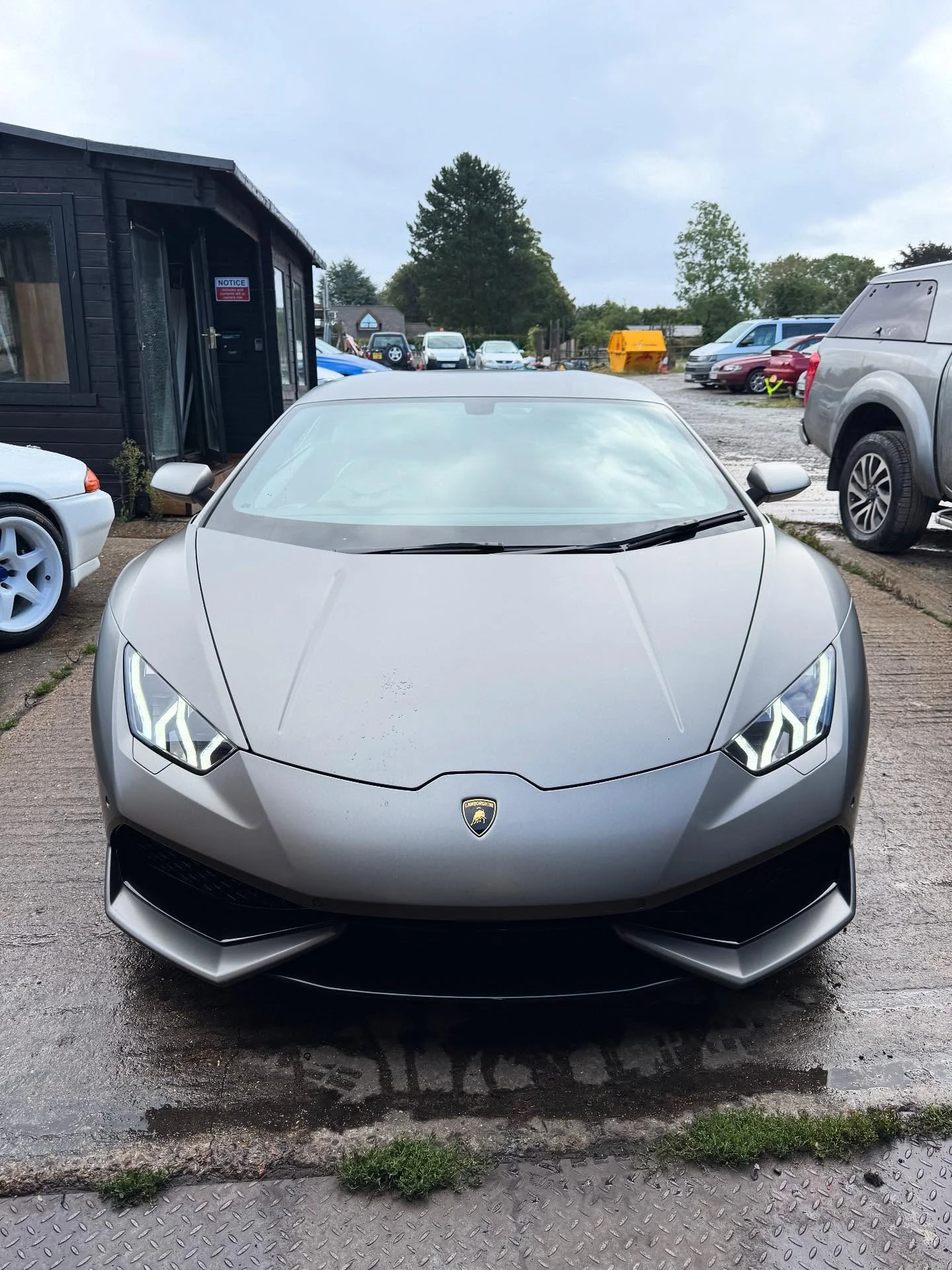 Front view of a silver Lamborghini with illuminated headlights parked on a concrete surface.