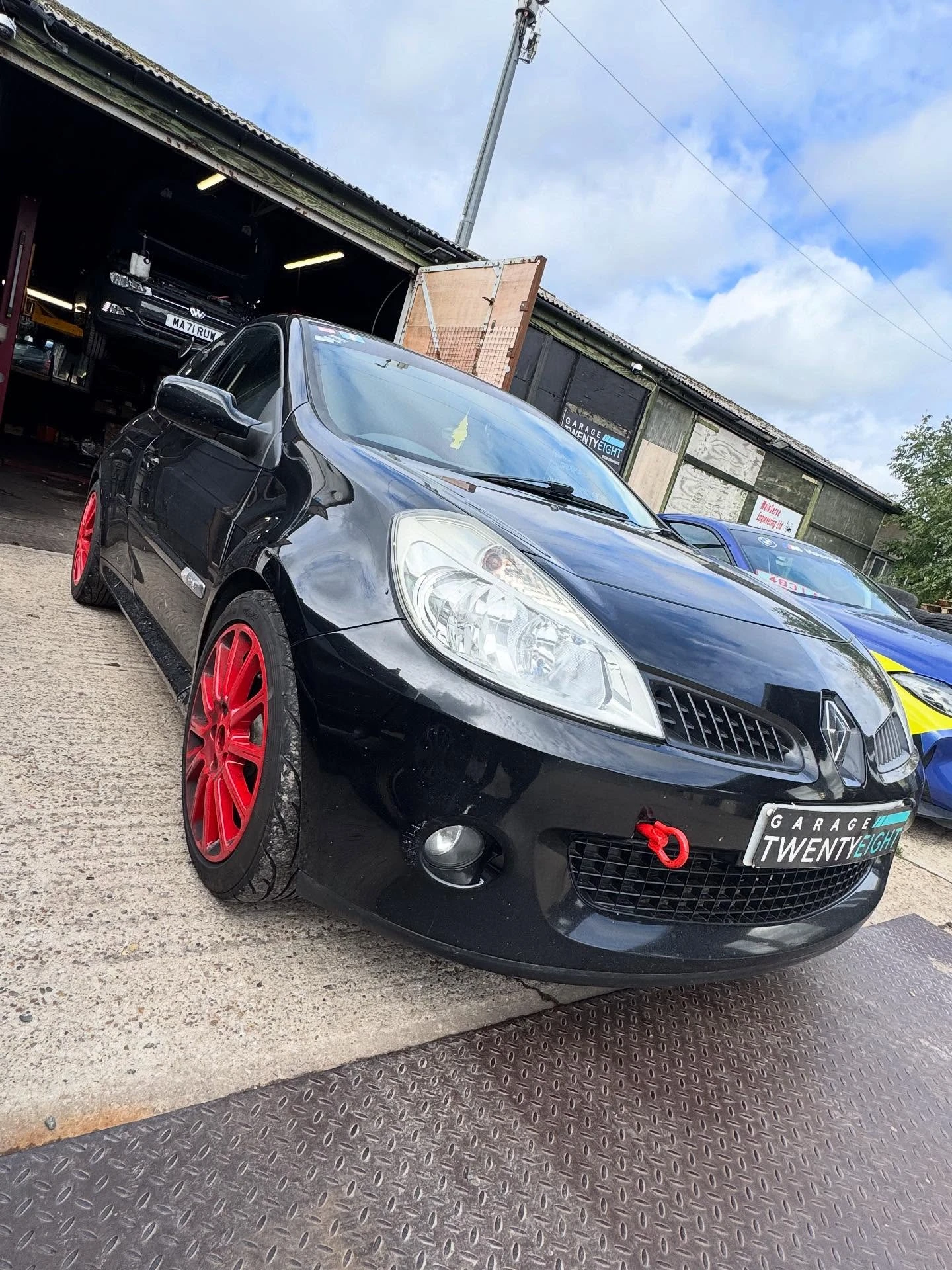 Black Renault car with red wheels parked outside a garage under cloudy sky.