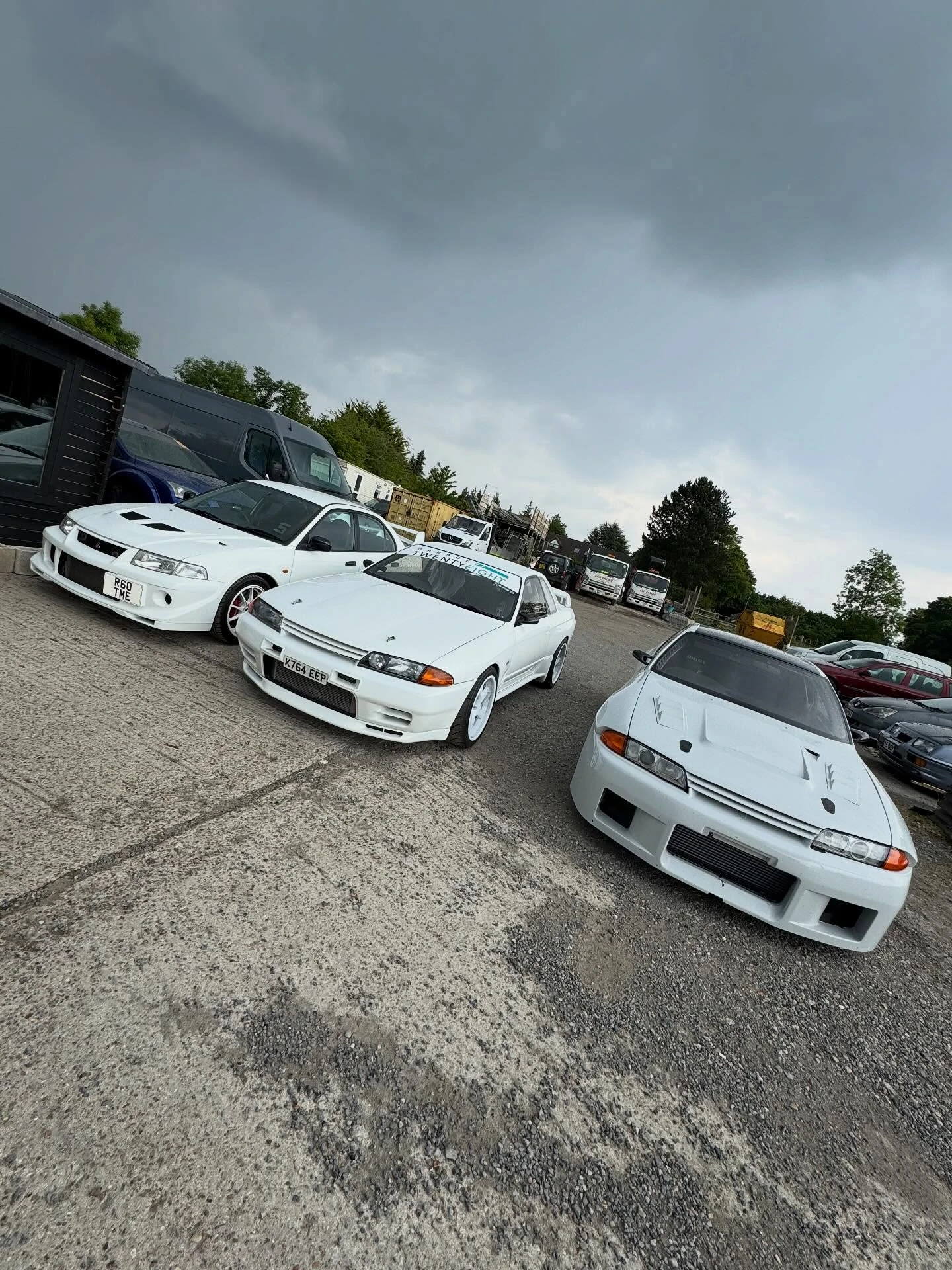 Three white sports cars parked outdoors on a gravel and concrete surface under cloudy sky.